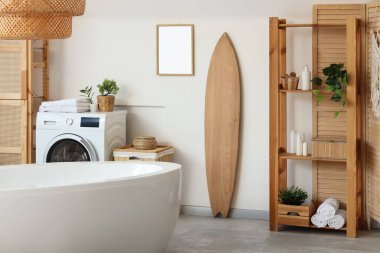 Küvet, çamaşır makinesi, ahşap mobilya ve sörf tahtasıyla aydınlık banyonun içi.