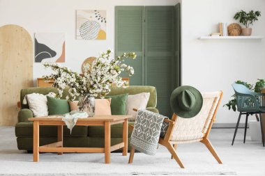 Yeşil kanepesi, koltuğu ve vazoda çiçekli dalları olan aydınlık oturma odasının içinde.