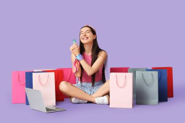 Kredi kartı ve dizüstü bilgisayarı olan mutlu genç bir kadın leylak arka planından alışveriş yapıyor.