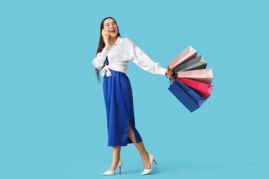 Alışveriş torbalarıyla mutlu genç bir kadın mavi arka planda cep telefonuyla konuşuyor.