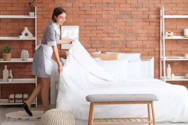 Genç oda hizmetçisi şık bir yatak odasında yatağı yapıyor.