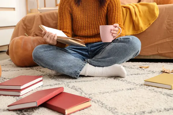 Elinde kahve fincanı olan kadın evde halıda kitap okuyor.