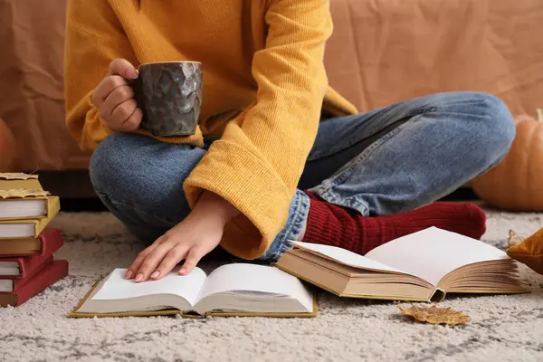 Elinde kahve fincanı ve kitaplarıyla evdeki halıda oturan bir kadın.