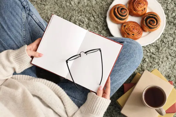 Boş kitaplı, gözlüklü ve kalçalı bir kadın halının üstünde oturuyor.