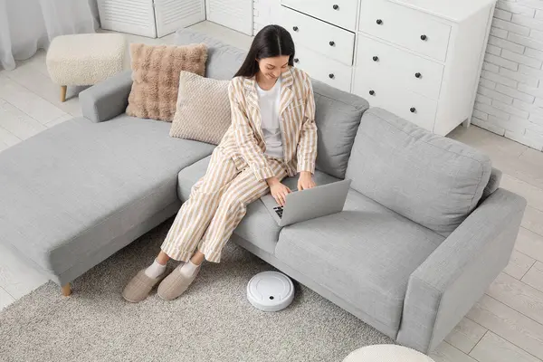 Pijamalı genç bir kadın evinde robot elektrikli süpürgeyle kanepede dizüstü bilgisayar kullanıyor.