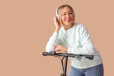 Tekme scooterlı ve kulaklıklı güzel olgun bir kadın bej arka planda müzik dinliyor.