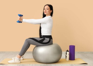 Spor yapan genç bir kadının portresi. Bej arka planda topa uygun halterlerle egzersiz yapıyor.