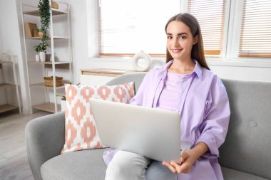 Genç ve güzel bir kadın dizüstü bilgisayarla sohbet ediyor ve evdeki kanepede oturuyor.