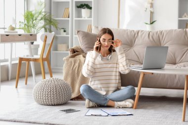 Laptop, tablet ve panolarla evde cep telefonuyla konuşan güzel genç bir kadın.