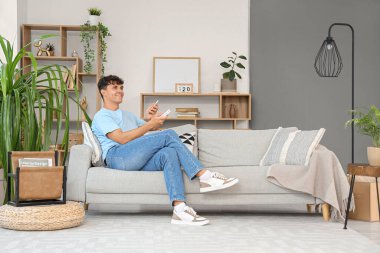 Cep telefonlu yakışıklı genç adam evdeki kanepede klimayı çeviriyor.