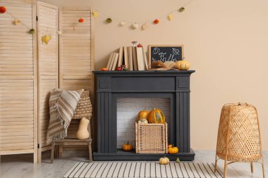 Bej duvarın yanındaki modern şöminede katlanan perdeli, kitaplı ve sonbahar dekorasyonlu şık bir oda.