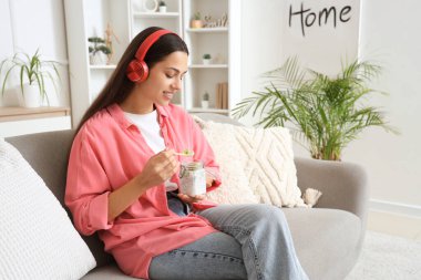 Kulaklıklı, Chia tohumu pudingli genç bir kadın evdeki kanepede oturuyor.