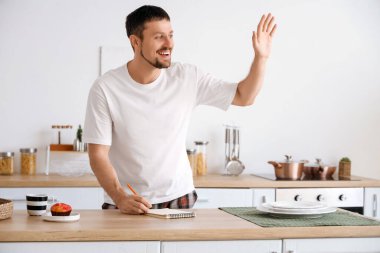 Pijamalı, not defterli, kahveli ve evde el sallayan mutlu genç adam.