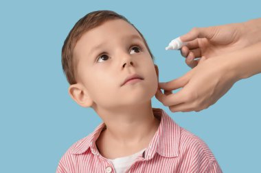 Mavi arka planda işitme sorunu olan küçük çocuğun kulağına damla damlatan kadın doktor, yakın plan.