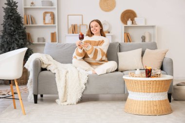 Sıcak süveter giymiş, elinde cam bardak sıcak şarapla evdeki kanepede oturan genç ve güzel bir kadın.