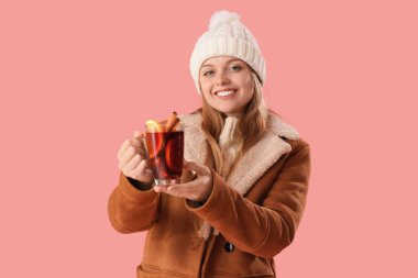 Pembe arka planda cam bardakta sıcak sıcak şarap ve kalın giysiler içinde güzel genç bir kadın.