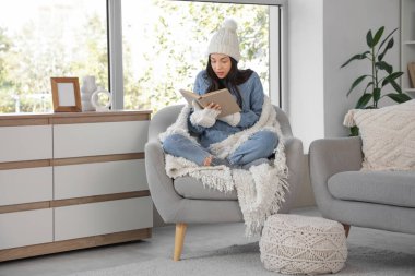 Evdeki koltukta ekose kitap okuyan dondurulmuş genç bir kadın.