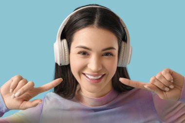 Güzel, genç, mutlu, kulaklıklı bir kadın mavi arka plandaki gülüşünü işaret ediyor.