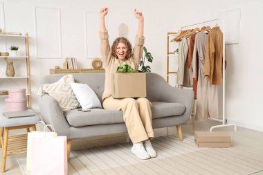 Evdeki koltukta yeni kıyafetlerle paket kutusunu açan mutlu genç kadın.