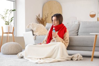 Evde dizüstü bilgisayar kullanarak öksürük şurubu içen hasta genç bir kadın.