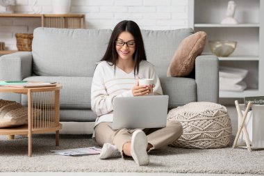 Oturma odasında dizüstü bilgisayar ve bir fincan kahveyle çalışan güzel genç bir kadın.