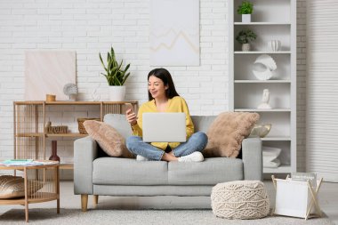 Oturma odasındaki kanepede oturan, cep telefonu ve dizüstü bilgisayarla çalışan güzel genç bir kadın.