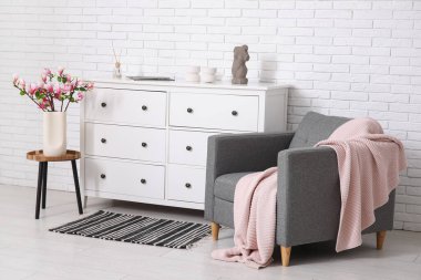 Çekmeceler, koltuk ve kahve masası. Odadaki beyaz tuğla duvarın yanındaki vazoda çiçek açan manolya dalları.