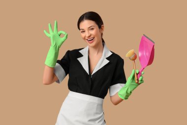 Lastik eldivenli, faraş ve fırçalı güzel oda hizmetçisi bej arka planda iyi bir jest gösteriyor.