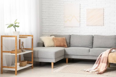 Beyaz tuğla duvarın yanında gri kanepesi ve raf ünitesi olan aydınlık oturma odasının içinde.