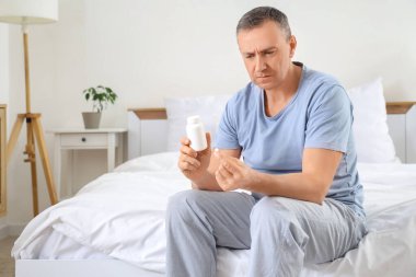 Elinde ilaç şişesi olan olgun bir adam yatak odasında oturuyor.