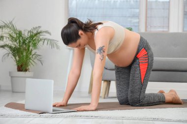 Bilgisayarlı hamile bir kadın evde yoga yapıyor.