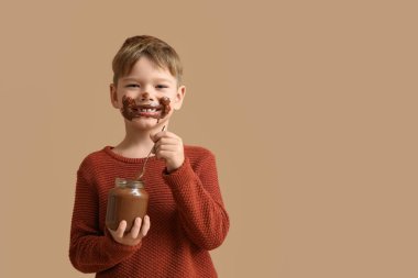 Yüzü lekelenmiş tatlı bir çocuk bej arka planda kavanozdan çikolata ezmesi yiyor.