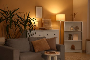 Oturma odasının içinde raf ünitesi, kanepe ve bej duvarın yanında parlayan lambalar var.