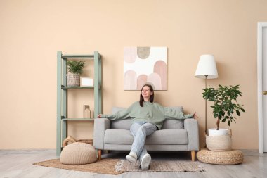 Oturma odasındaki bej duvarın yanındaki kanepede oturan mutlu genç kadın.