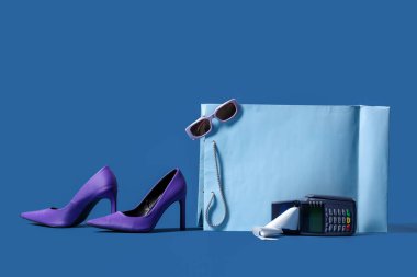 Şık güneş gözlüklü alışveriş çantası, yüksek topuklu ayakkabı ve mavi arka planda ödeme terminali.