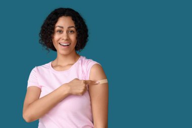 Genç Afro-Amerikalı kadın mavi arka plandaki aşıdan sonra uygulamalı tıbbi yamaya işaret ediyor.