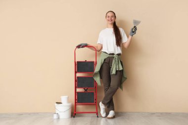 Bej duvarın yanında elinde oyuncak bıçak ve merdiven olan genç bir kadın.