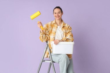Leylak arka planına karşı merdiven üzerinde boya silindiri ve kova taşıyan genç bir kadın.