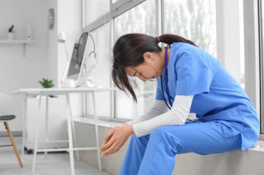 Klinikte pencere kenarında oturan üzgün Asyalı kadın doktor.