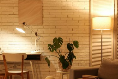 Gece oturma odasının içinde koltuk, katlanan ekran, parlayan lambalar ve beyaz tuğla duvarın yanındaki işyeri.