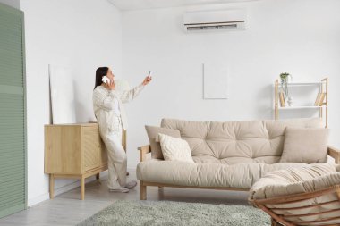 Oturma odasında klimayı açarken cep telefonuyla konuşan genç bir kadın.