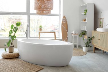 Küveti, büyük penceresi, ev bitkileri ve sörf tahtasıyla aydınlık banyonun içi.