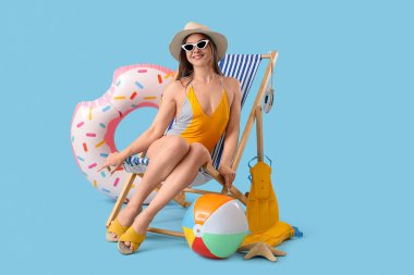 Güzel bir kadın mavi arka planda plaj aksesuarlarıyla güverte koltuğunda oturuyor.