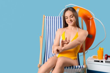 Güzel bir kadın mavi arka planda iskemlede güneş kremi sürüyor.