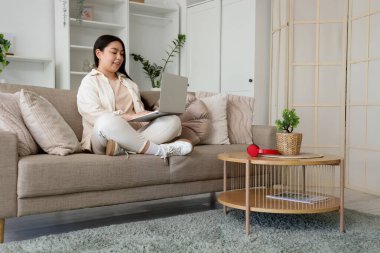 Evdeki kanepede dizüstü bilgisayar kullanan gülümseyen genç kadın.