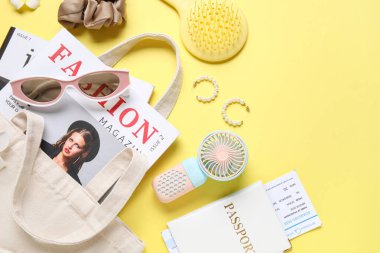 İçinde kadın aksesuarları, moda dergileri, pasaport, bilet ve sarı arka planda mini fan olan bir çanta.