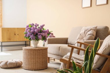 Oturma odasının içinde kanepe, koltuk ve vazoda leylak çiçekleri var.