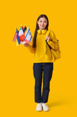 Farklı ülkelerin bayrakları ve sarı arka planda sırt çantasıyla genç bir Asyalı kadın.