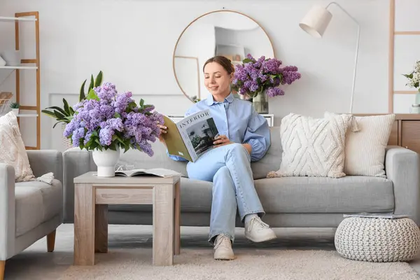 Oturma odasında kanepede ve vazoda leylak çiçekleri olan dergi okuyan genç bir kadın.