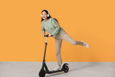 Kulaklıklı güzel bir Asyalı kadın sarı duvarın yanında modern elektrikli scooter kullanıyor.